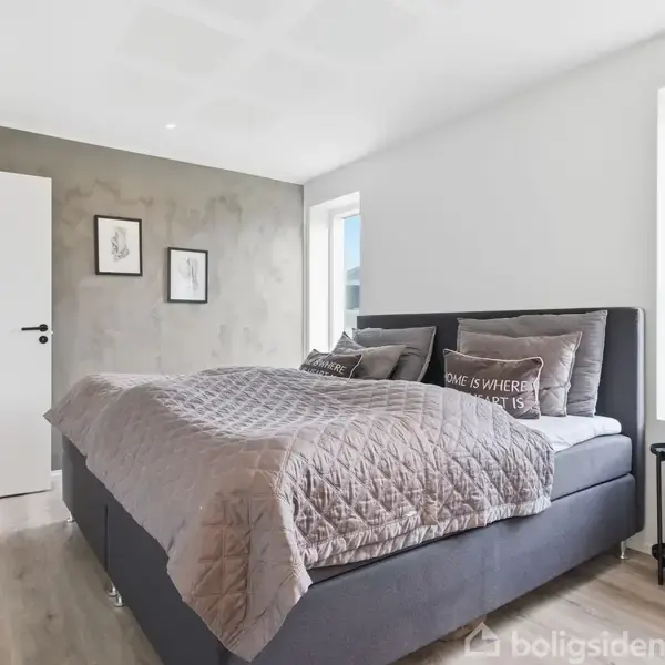 En dobbeltseng står i et lyst soveværelse, dækket af en quiltet grå dyne. Ved siden af er et lille bord med en plante. Væggen har tre indrammede billeder. En pude har teksten "HOME IS WHERE THE HEART IS".