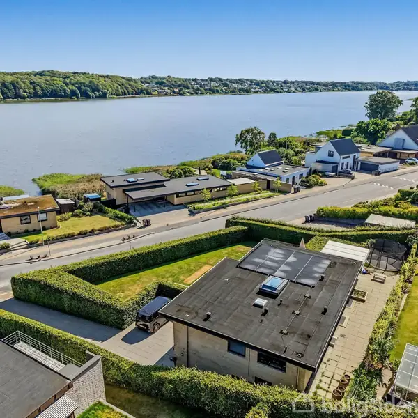 Hus med fladt tag og have ligger ved en vej, som løber langs en bred sø. Omkringliggende huse og frodig vegetation omkranser området under en klar blå himmel.