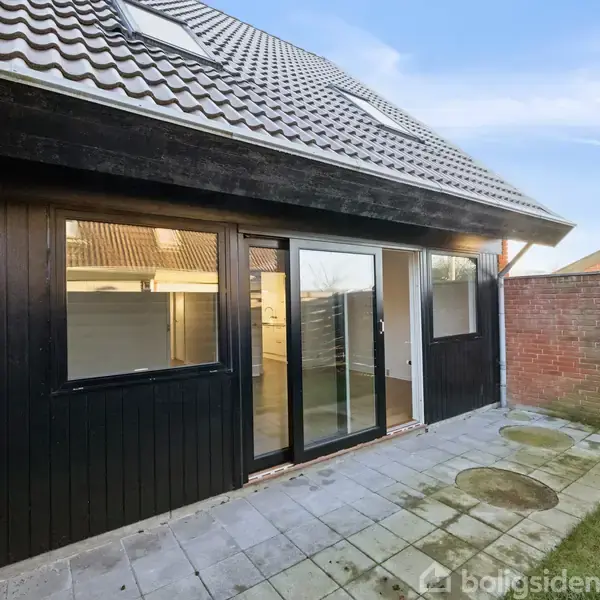 Et sortmalet hus med store vinduer og en glasdør står i ro på en flisebelagt terrasse. Omkring ses en murstensvæg og en træhegn, under en blå himmel.