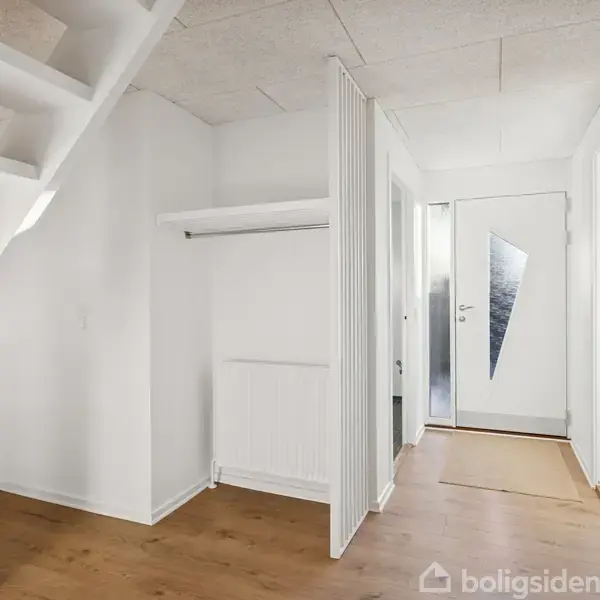 Trappe i træ fører opad i moderne, lyst entrérum. Rummet har hvide vægge, trægulv og en glasdør med et unikt, trekantet mønster.