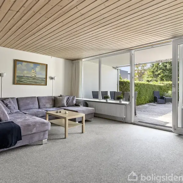 Grå hjørnesofa står i en lys stue med træloft. Et kaffebord er centralt placeret. Store vinduer og glasdør giver udsigt til en terrasse med grønne buske udenfor.