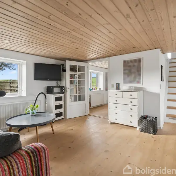 Stue med grå sofa, rundt sofabord og farverig stribet pude. Træloft og gulv. Dør til venstre åbner mod spisestue. Fritstående kommode ved trappe. Væggene er hvide.
