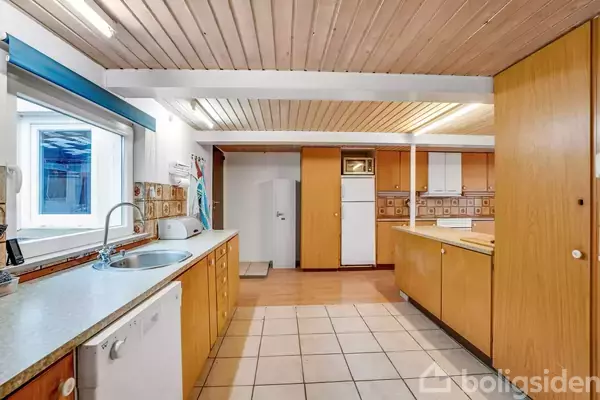 Et køkken med træloft, brune overskabe, og lyse fliser på gulvet. Et opvaskemaskine ved vasken, vindue til venstre, og køleskab samt skabe i baggrunden.