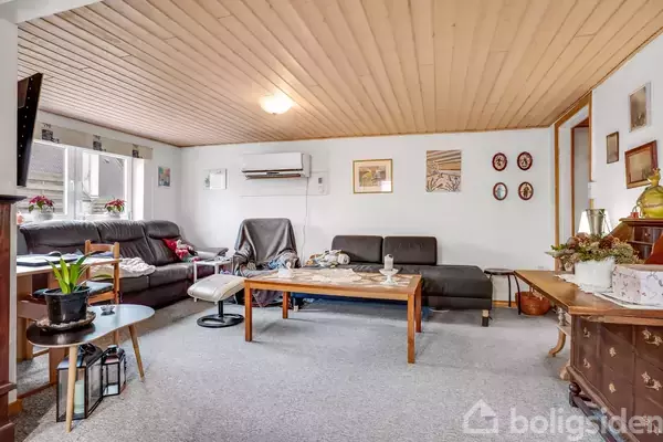 En stue med sorte lædersofaer står ved siden af et stort træbord. Der er en aircondition på væggen, og rummet er dekoreret med billeder og planter.