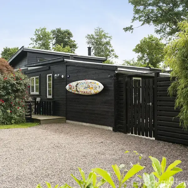 Et sort træhus står ved siden af en grusvej omgivet af buske og træer. På husets væg hænger et surfbræt med teksten "Casa Chalet Walk in Welcome".