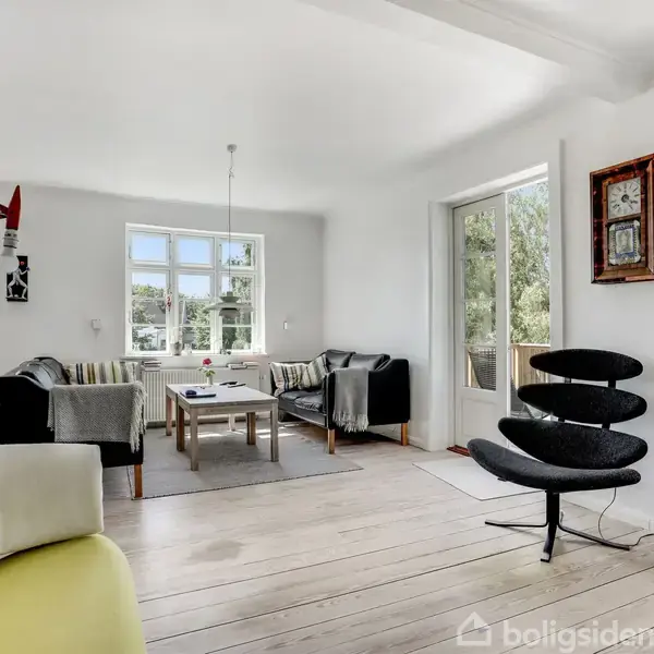 En stue med moderne møbler, hvor to sofaer omkranser et sofabord. Store vinduer og en glasdør lukker naturligt lys ind. Enkelte kunstværker er ophængt på væggene.