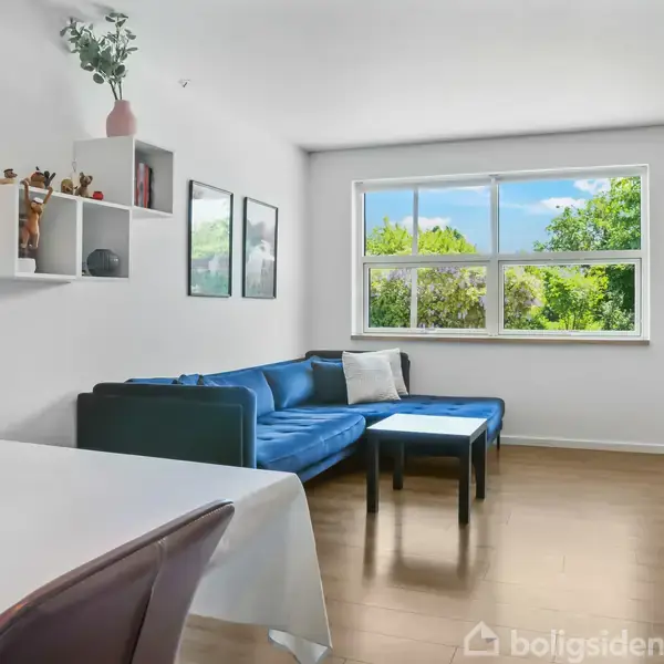 Blåt sofahjørne med sofabord i stue. Ved siden af er et spisebord med stol. Vindue viser grønt landskab. Væggen har hylder og billeder. Lampe hænger til højre.