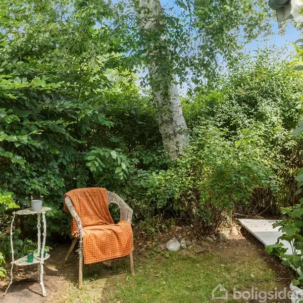 En stol med orange plaid står stille i en frodig have ved siden af et lille bord. Omgivet af grønne buske, træer og blomstrende planter nær et grønt hus.