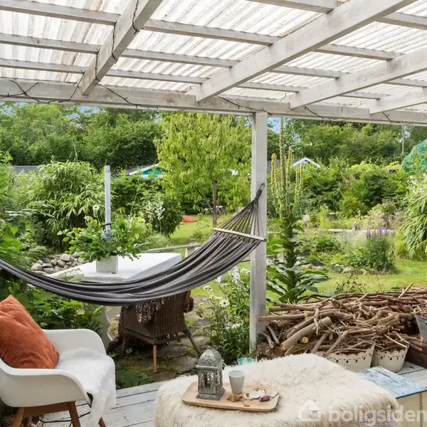 Hængekøje hænger under en overdækket terrasse omgivet af grønt og træstubbe. En stol med en pude står ved siden af et bord med lys og kopper.