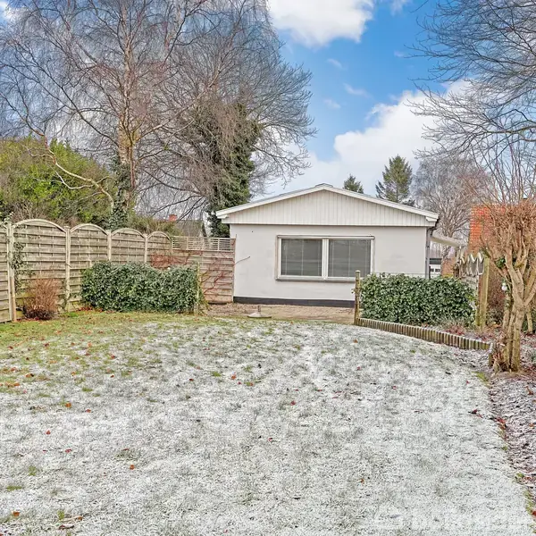 Et lille, hvidt hus står for enden af en snedækket græsplæne omgivet af buske og træer. En trærækværk danner hegn i haven under en blå, let skyet himmel.