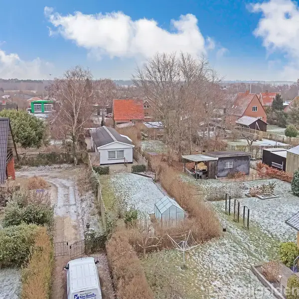 Huse med snedækkede tage står stille i en villakvarter. Haver er let dækket af sne, med hegn og tidlig morgenlys i baggrunden under en blå himmel.