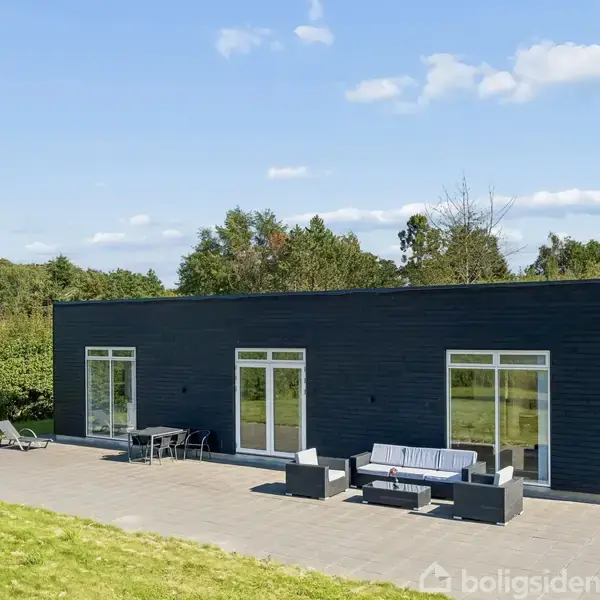 En moderne, sortmalet étplansvilla står stille på en flisebelagt terrasse. Møbler med parasol og sofa er placeret foran store vinduer. Græs og træer omgiver huset under en blå himmel.