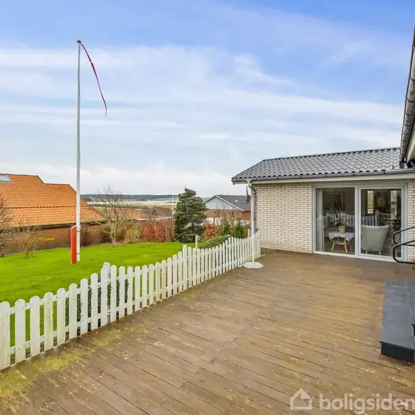 En træterrasse med potteplanter grænser op til et hvidt hegn. I baggrunden ses en velplejet have med et flagstang og udsigt over et hus og en vandkrop.