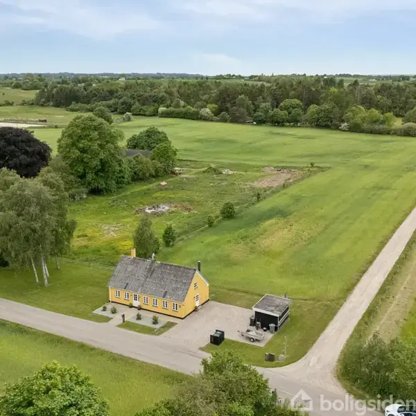 Gult hus med sort tag står alene midt i grønne marker og træer, ved siden af en landevej.