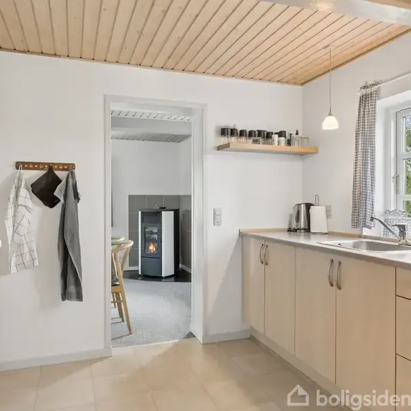 Et lyst og moderne køkken med træloft og lyst trægulv. Køkkenet er udstyret med en vask og en række hylder. En dør fører til et rum med en pejs.