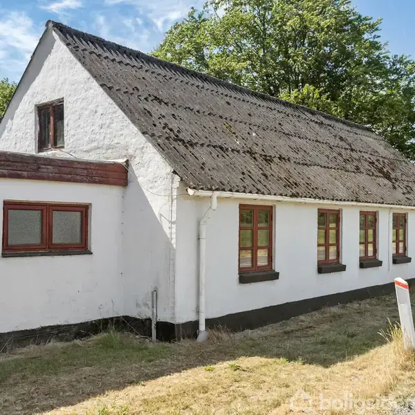 Et hvidt, stråtækt hus står stille ved en vej, omgivet af grønne træer. Flere vinduer langs facaden giver indtryk af en landlig idyl.