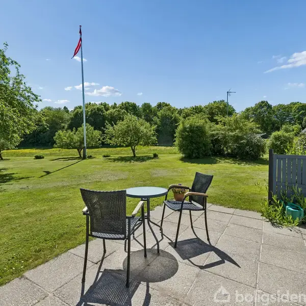 To havestole og et bord står på en terrasse, omgivet af en stor græsplæne med træer. En flagstang med flag er synlig i haven under en klar, blå himmel.