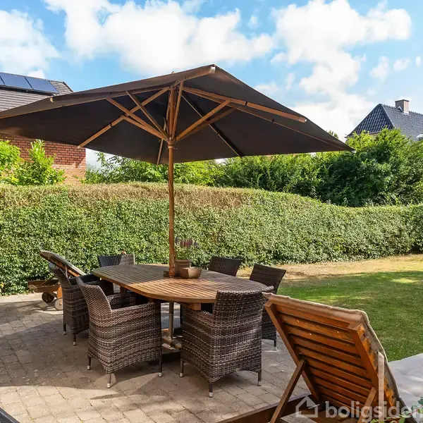 Et havebord med seks stole står på en flisebelagt terrasse under en stor parasol. I baggrunden er der en velplejet have med grøn hæk og enkelte træer.