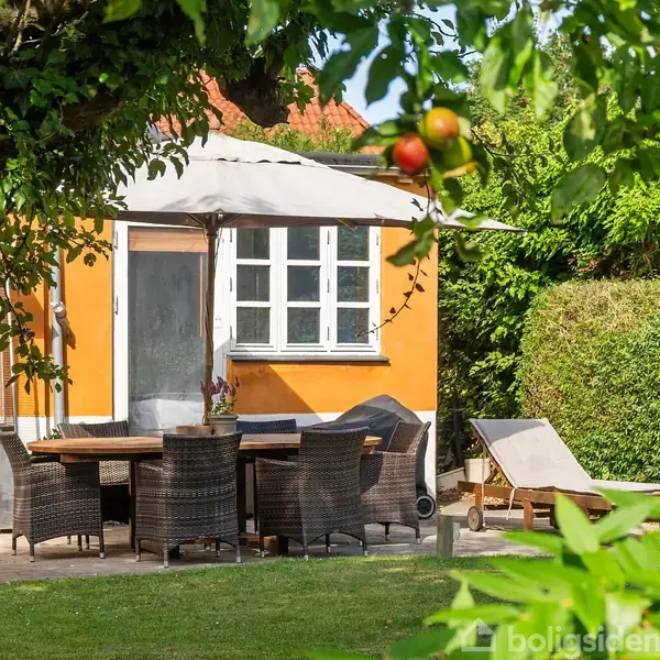 Et gult hus med hvide vinduer står i en have. Foran huset er et bord med stole og en parasol. Græs, buske og frugttræer omgiver området.