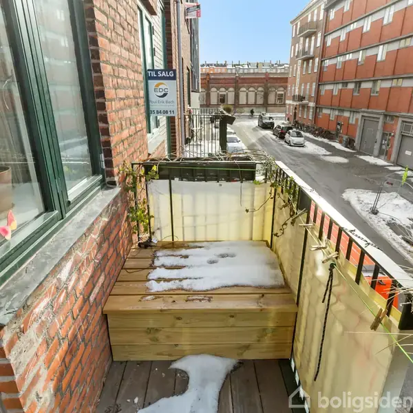 Balkon dækket med sne, omgivet af murstensvægge, med udsigt til en gade. Et "Til salg" skilt fra EDC hænger på rækværket.