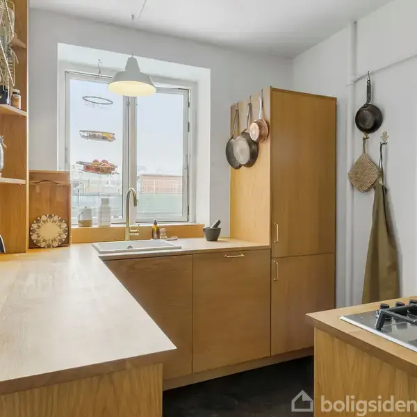 Køkken med gaskomfur og træskabsplads. Aktion: Sollys strømmer ind gennem vinduet. Kontekst: Køkkenredskaber hænger på væggene; hænder fyldes med lys fra hængelampe.