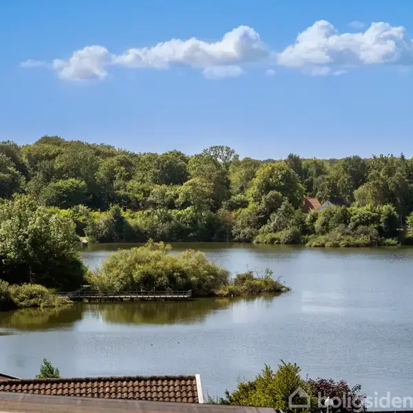 En sø strækker sig roligt i forgrunden med frodig, grøn skov omkring. Under en blå himmel kan husets tag fra venstre side ses i nederste kant.