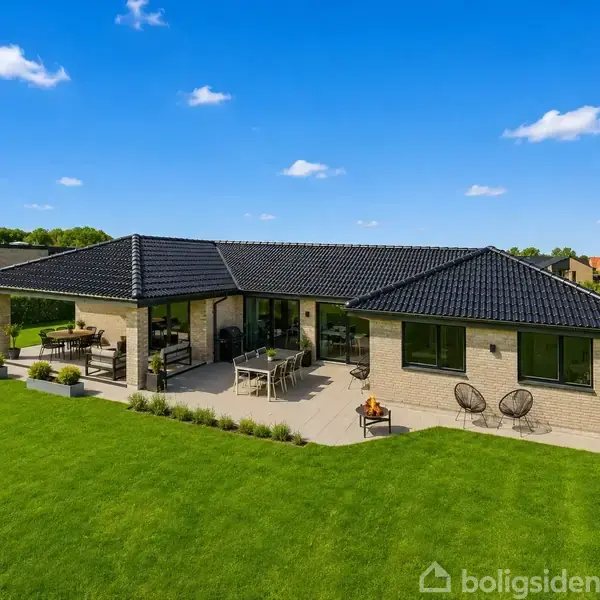 En moderne bungalow med sort tegltag står på en velplejet græsplæne. Huset har en stor terrasse med havemøbler og er omgivet af grønne buske under en klar, blå himmel.