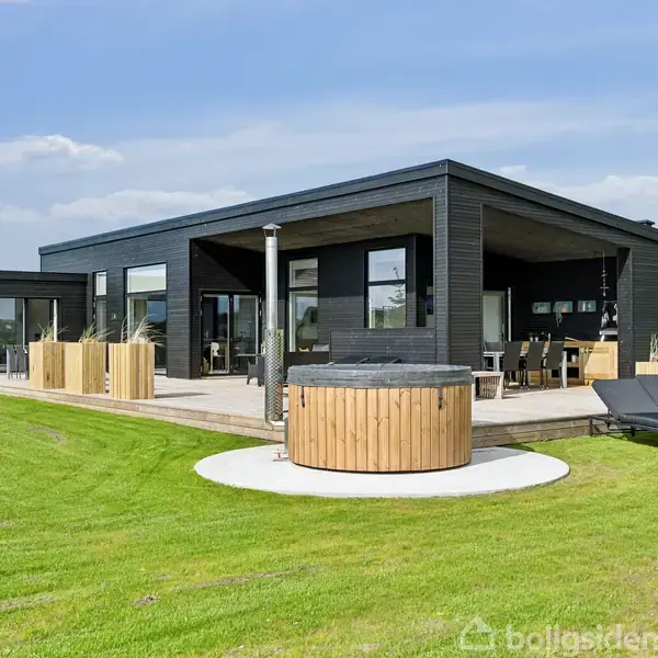 En moderne, sortmalet bungalow med store vinduer og en træterrasse med spa. Solsenge står ved siden af, omgivet af en velplejet græsplæne under en klar himmel.