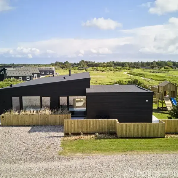 Sort, moderne hus står stille omgivet af grønne marker og et grusområde i forgrunden. Et legehus står i baggrunden, mens himlen er let overskyet.