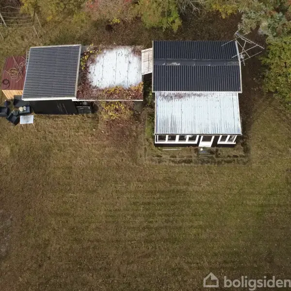 To huse med metal- og tjæretage står side om side i en græsplæne, omgivet af træer. Ved siden af står et hvidt køretøj.