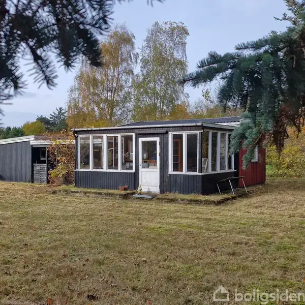 Et sort træhus med mange vinduer står på en græsplæne. Ved siden af er der et skur. Omgivet af træer i efterårsfarver.