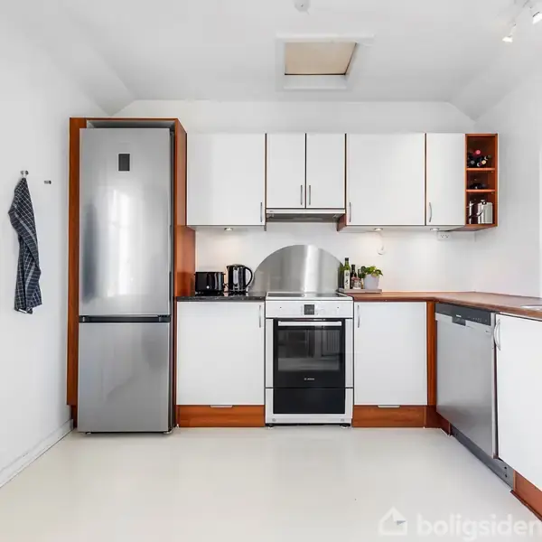 Køkken med hvide skabe og stålapparater; køleskab og ovn omgivet af trækanter. Vægophængt viskestykke. Vindue til højre med plante. Rummeligt og lyst interiør.