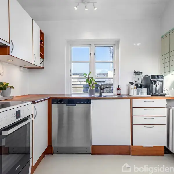 Køkken med hvidt interiør og trædetaljer. Opvaskemaskine og kaffemaskine står på en lys bordplade. En plante og køkkenredskaber er ved et vindue med dagslys. Radiator til højre.