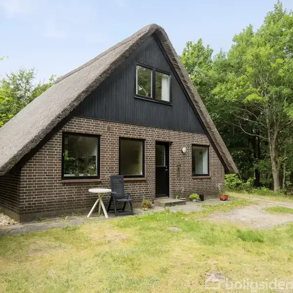 Hus med stråtag og murstensvægge står stille i en grøn have omgivet af træer. Foran huset er der en lille terrasse med et bord og en stol.