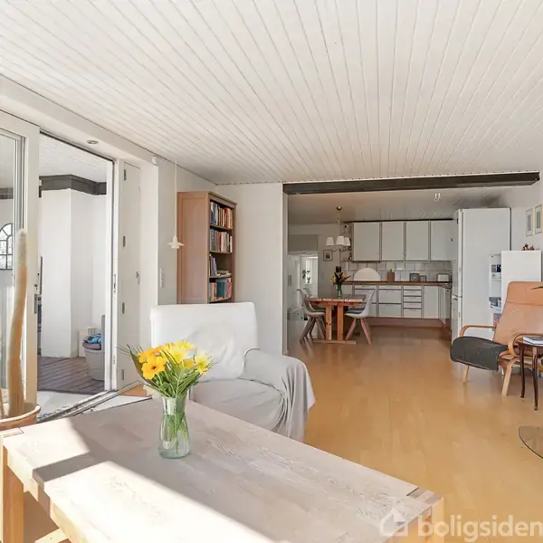 Stue med sofa og sofabord med gule blomster, træbrændeovn i hjørnet. Åben adgang til køkkenet i baggrunden. Lyst og rummeligt miljø med trægulve og hvide vægge.