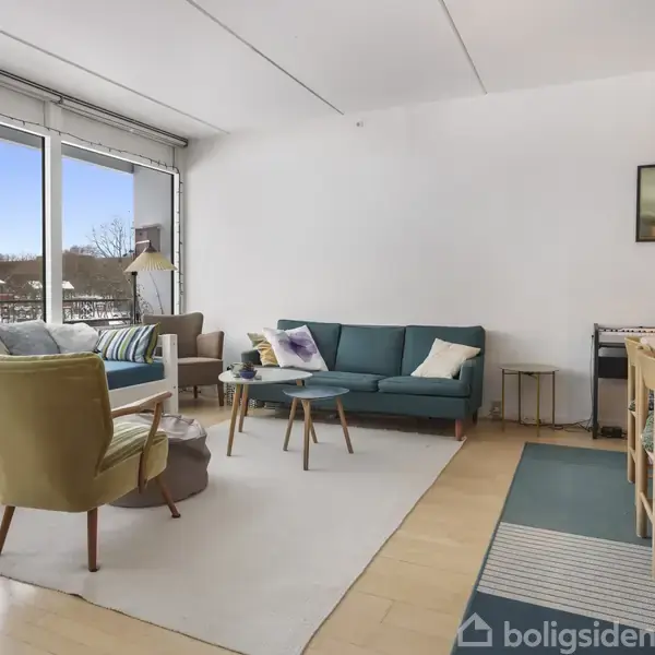 En stue med lyse træborde og -stole, en sofa ved et vindue samt en lænestol. Udendørs ses sneklædt landskab gennem store vinduer. Vægdekoration og lamper tilsætter hyggelig stemning.