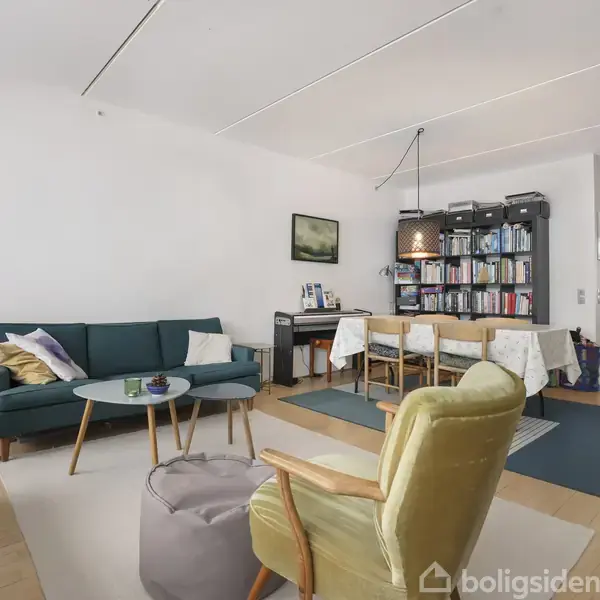 Sofa står i stue med bord og puder, mens reoler fyldt med bøger pryder væggen bag spisebordet. Rummet har lyst trægulv og moderne indretning.