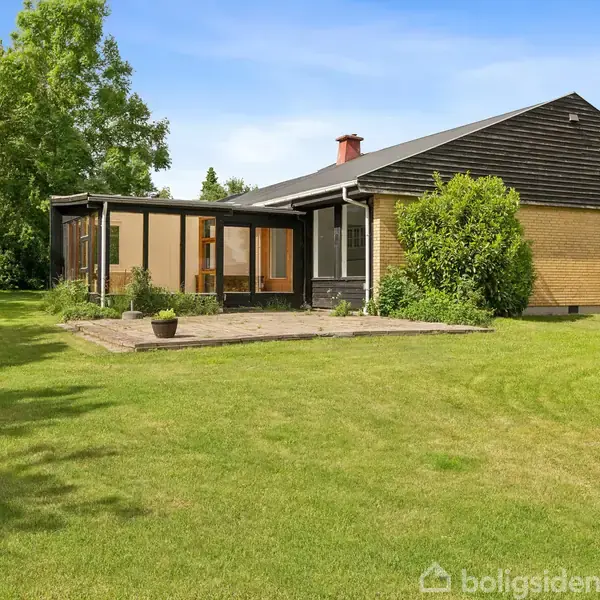 Et én-etagers hus med gulstensvægge og skråt tag står stille. Foran huset er en veranda med store vinduer, omgivet af en velplejet græsplæne i et åbent og solrigt område.