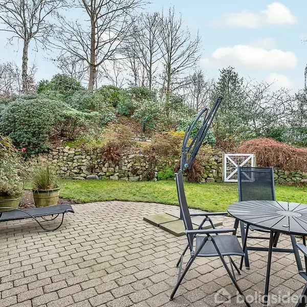 Et rundt havebord med stole står på en flisebelagt terrasse. I baggrunden er der en græsplæne og en stensætning med buske og træer, der giver et hyggeligt udendørsmiljø.