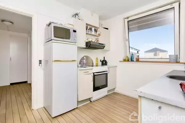 Køkken med køleskab, mikrobølgeovn, komfur og vask. Trægulv og stort vindue med udsigt. Hylder med køkkenartikler hænger over vasken. Lyse vægge skaber en ren, minimalistisk atmosfære.