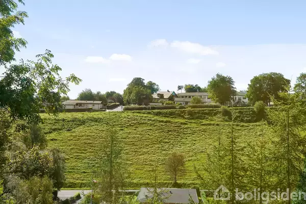 En grøn bakke strækker sig forbi en landevej, hvor flere huse og træer ses i baggrunden under en blå himmel med få skyer.