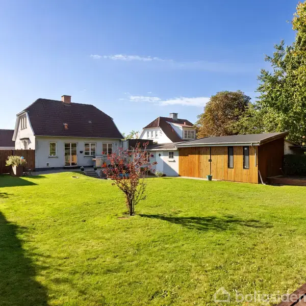 Et hvidt hus står i en grøn have med en rød busk i midten. Til højre ses en træbygning. Solrigt vejr og en klar blå himmel omfavner scenen.