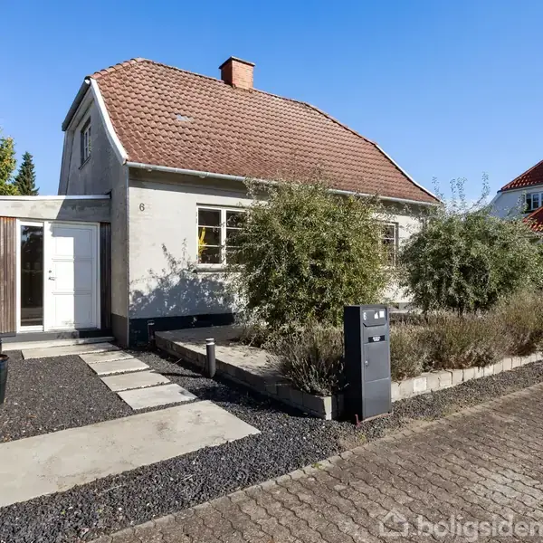 En hvid murstensvilla med brunrødt tag har en havelåge og fliser, omgivet af buske. Solrig dag med blå himmel. Adresse "8" er synlig ved indgangspartiet.