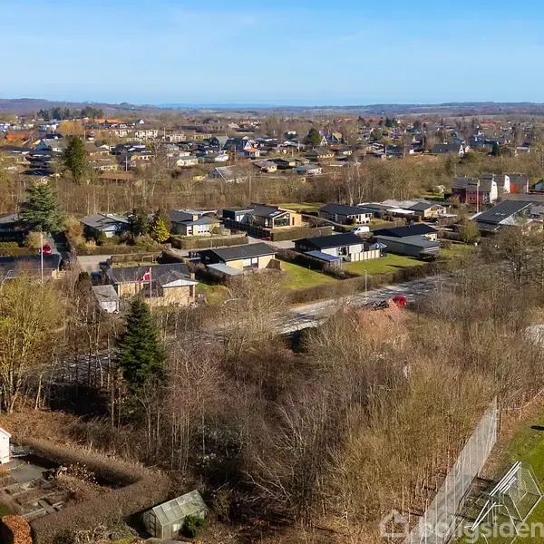 Et boligområde set fra oven viser huse tæt på hinanden med frodige haver og træer rundt omkring. I baggrunden strækker byen sig mod horisonten under en klar blå himmel.