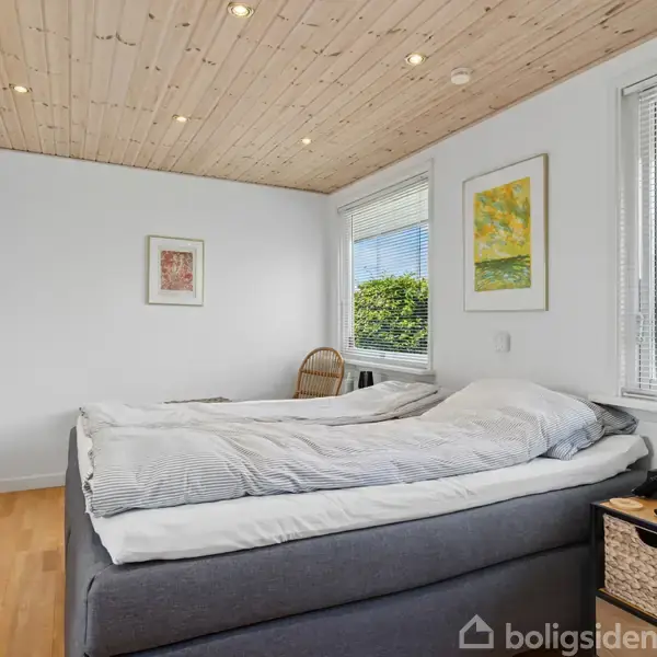 En seng med gråt betræk står i et lyst soveværelse. To billeder hænger på de hvide vægge. Vinduet viser en grøn have. En kurvestol står i hjørnet.