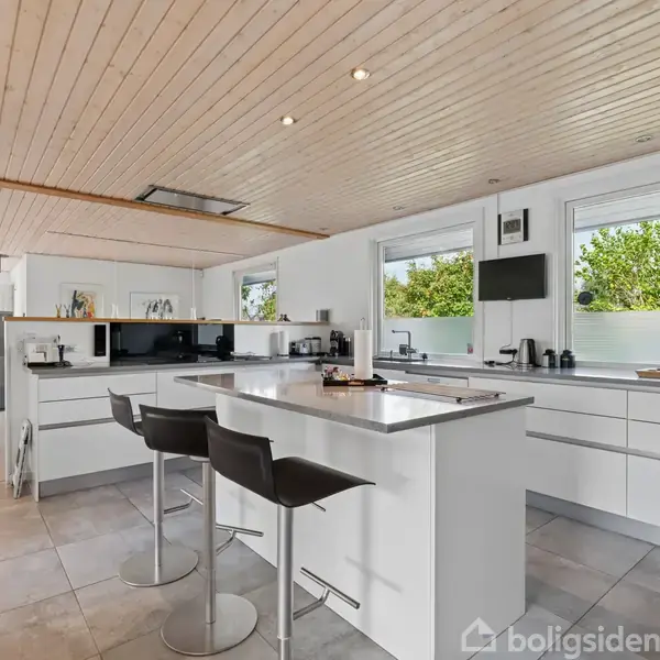 Et moderne køkken med en stor ø. To barstole står ved øen. Rummet har lyse trælofter, hvide skabe og store vinduer med udsigt til grønne træer.