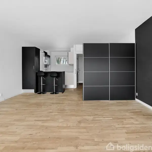 Rummelig stue med lyst trægulv, opdelt af en stor, sort skillevæg. Et køkkenområde med to barstole er synligt i baggrunden, omgivet af hvide vægge og sort interiør.