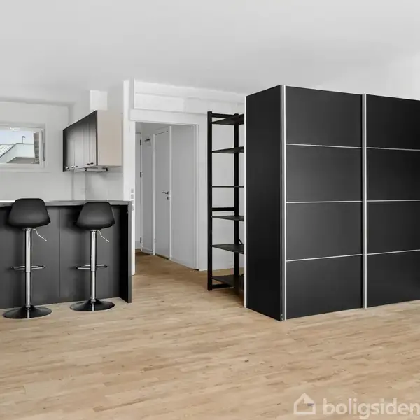 En moderne lejlighed med et åbent opholdsområde. To sorte barstole står ved en køkkenbar, og sorte skabe fylder væggen. Lyse trægulve i en minimalistisk indretning.