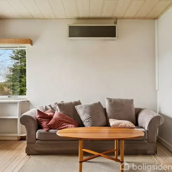 Sofa med puder står foran et lille rundt bord i et lyst rum med trægulv, store vinduer til venstre og en døråbning til højre. En radiator er placeret over sofaen.
