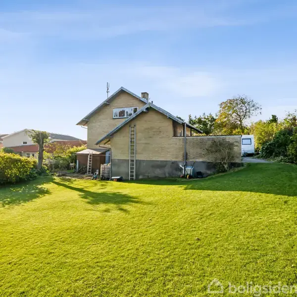 Hus omgivet af en grøn græsplæne og træer; en stige læner sig mod husets mur. Området er solrigt med en klar blå himmel.